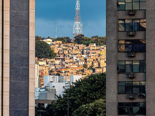 Um morro densamente ocupado por casas de alvenaria e telhados de cerâmica é visto ao longe, enquadrado por dois grandes edifícios em primeiro plano. No topo do morro, uma alta torre de transmissão branca e vermelha se destaca contra um céu de cor azul-escuro e nublado.