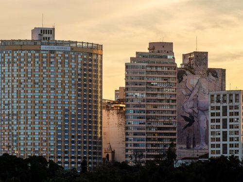 Dois grandes edifícios se destacam no centro da imagem. O da esquerda é arredondado, com fachada de pastilhas amarelas. No prédio da direita, há um grande mural de arte urbana com figuras humanas em tons de cinza. A cena é capturada durante o pôr do sol, com um céu de cor amarelada ao fundo.