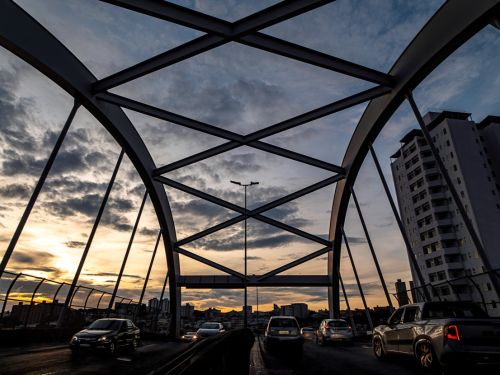 Vista de baixo para cima da estrutura metálica em arco do viaduto, que forma uma moldura geométrica contra o céu. Carros trafegam em ambos os sentidos. À direita, um prédio alto e branco. O céu ao fundo tem nuvens escuras e tons alaranjados do pôr do sol no horizonte.