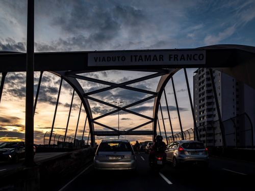 Vista a partir de um veículo no trânsito, olhando através da estrutura do viaduto. Uma viga horizontal no topo tem a inscrição "VIADUTO ITAMAR FRANCO". Carros e uma motocicleta estão parados à frente. Ao fundo, o céu exibe cores do final de tarde, com nuvens e tons de azul e laranja.