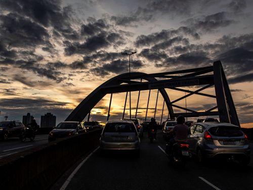 A silhueta do arco do viaduto se destaca contra o céu de fim de tarde. Várias pistas de trânsito estão congestionadas com carros e motocicletas. A luz do sol poente, de cor amarela, brilha sob o arco. O céu está coberto por nuvens escuras e texturizadas.