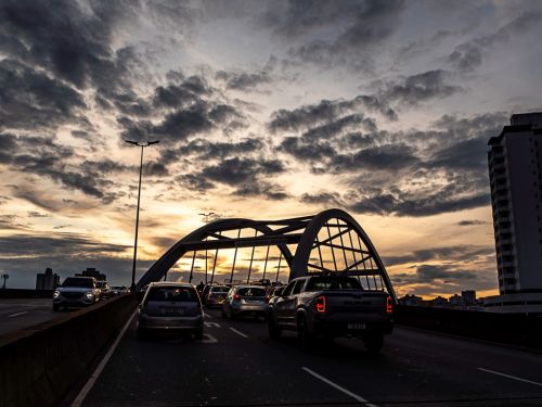 Carros em fila sobre o viaduto ao entardecer. A estrutura em arco do viaduto está à frente, em silhueta. À direita, um prédio alto. O céu está coberto por nuvens escuras, mas o horizonte é iluminado por uma faixa de luz amarela do sol poente, que passa por baixo do arco da ponte.
