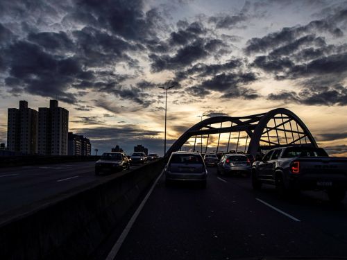 Vista do trânsito sobre o viaduto, com a maior parte da cena em silhueta escura. O arco da ponte se destaca contra o céu. Prédios à esquerda também estão em silhueta. O céu está repleto de nuvens escuras, com uma faixa de luz amarela do crepúsculo visível no horizonte.