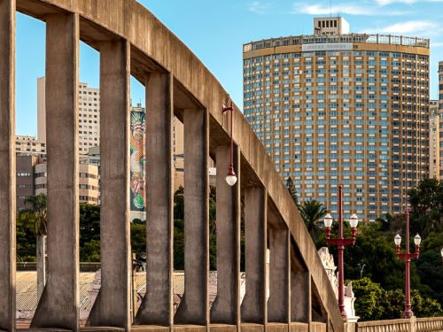 Visão em diagonal de um dos arcos de concreto do viaduto, com seus pilares verticais em primeiro plano. Através dos pilares, vê-se um grande edifício com fachada de pastilhas amarelas e janelas quadradas. Ao fundo, um mural colorido na empena de outro prédio e postes de iluminação vermelhos.