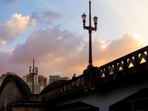 A silhueta escura do Viaduto Santa Tereza é vista em contra-luz durante o pôr do sol. Um poste de iluminação ornamental de cor vinho se destaca no centro. O céu tem nuvens com tons de rosa e laranja. Ao fundo, a silhueta de prédios e o arco de concreto do viaduto à esquerda.