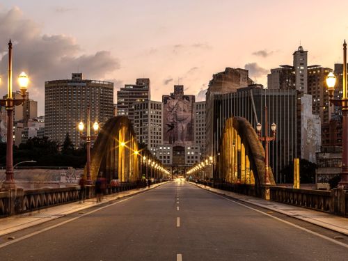 Vista frontal do viaduto ao anoitecer. A pista de asfalto vazia leva o olhar ao centro da cidade. Os postes de iluminação de ambos os lados estão acesos, com uma luz amarela quente. Ao fundo, o grande mural em um prédio e os arcos do viaduto se destacam contra o céu de cor alaranjada.