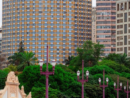 Vista lateral do viaduto, mostrando a pista de asfalto e uma fileira de postes de iluminação de cor vinho. Logo abaixo, uma densa vegetação verde. Ao fundo, um grande edifício de formato arredondado, com fachada de pastilhas laranjas e brancas, se destaca contra o céu nublado.