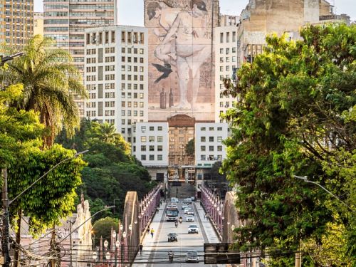 Fotografia de um ponto elevado, olhando para a descida de uma rua em direção ao Viaduto Santa Tereza. Carros estão estacionados nas laterais. O viaduto, com seus arcos e postes, atravessa a parte central da imagem, levando a um corredor de prédios onde se destaca um grande mural de arte urbana.