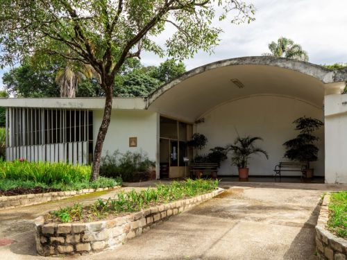 Prédio branco de entrada no zoológico, com arco curvo, janelas enfileiradas, bancos e vasos de plantas. Na frente, jardins com flores e arbustos cercados por pedras, ao lado de árvores altas.