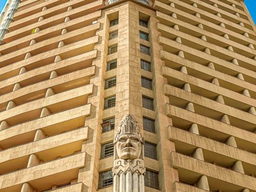 A vista de baixo para cima da fachada principal do Edifício Acaiaca. O prédio tem um tom bege, com andares que se projetam para fora, formando varandas em toda a sua extensão. No centro da fachada, uma escultura gigante de uma cabeça indígena com um cocar se destaca, apoiada sobre uma coluna que se estende para baixo.