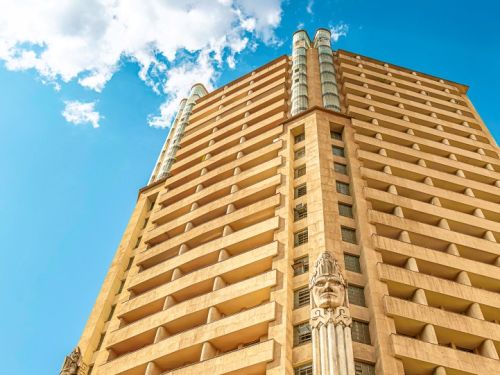 A vista de baixo para cima da fachada principal do Edifício Acaiaca. O prédio tem um tom bege, com andares que se projetam para fora, formando varandas em toda a sua extensão. No centro da fachada, uma escultura gigante de uma cabeça indígena com um cocar se destaca, apoiada sobre uma coluna que se estende para baixo.