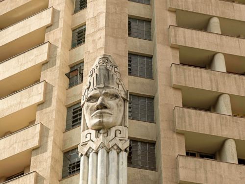Um close na fachada do Edifício Acaiaca, focando na icônica escultura de uma cabeça indígena. A escultura, que é o ponto focal da foto, está posicionada em uma coluna vertical de cor clara. Em ambos os lados da coluna, há partes do edifício. À esquerda, uma seção com janelas escuras e à direita, outra seção com várias varandas. A fachada do prédio tem uma cor bege e o design dos andares apresenta linhas retas e horizontais.