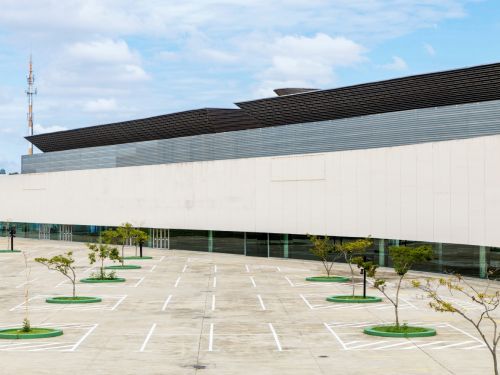 Fachada lateral de um edifício grande, de cor clara, com linhas retas e janelas contínuas na base. Na parte superior, há uma faixa metálica cinza e, acima dela, estruturas inclinadas de telhado escuro. Em frente ao prédio, há um amplo estacionamento de concreto, com várias vagas demarcadas em linhas brancas. O espaço conta com canteiros circulares pintados de verde, nos quais estão plantadas árvores jovens distribuídas em fileiras. No fundo, à esquerda, é visível uma torre metálica de telecomunicação.