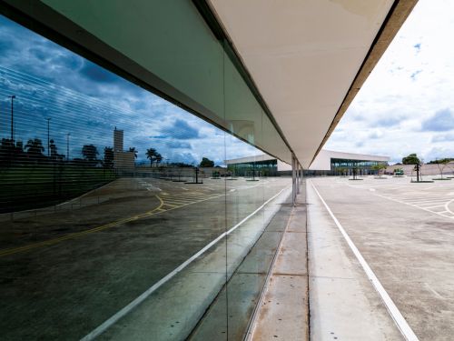 A imagem mostra uma área externa ampla, pavimentada em concreto, com marcações brancas e amarelas no chão que indicam vagas de estacionamento e faixas de circulação. À direita, vê-se a fachada envidraçada de um edifício moderno, com cobertura em linha reta. O vidro reflete parte da paisagem, incluindo árvores, postes de iluminação e o céu nublado. Ao fundo, há outro bloco do edifício, também de fachada envidraçada, além de árvores plantadas em canteiros espaçados ao longo do estacionamento.