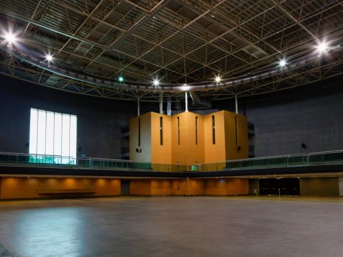 A imagem mostra o interior de um pavilhão de eventos. O espaço é amplo, com piso liso de concreto cinza e pé-direito alto. No centro do fundo do ambiente há uma estrutura de madeira clara, de formato retangular, com aberturas verticais estreitas. Acima, o teto metálico expõe vigas, tubulações e refletores circulares distribuídos ao redor. À esquerda, há uma grande janela vertical composta por painéis de vidro que permitem a entrada de luz natural. Nas laterais, há um mezanino com guarda-corpo de vidro, e na