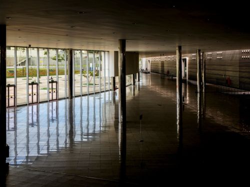 A imagem mostra um amplo hall interno com piso de revestimento brilhante em tons escuros, sustentado por colunas cilíndricas metálicas. À esquerda, há uma extensa parede de vidro que permite a entrada de luz natural e a visão da área externa, onde se vê calçada, gramado e algumas árvores. No fundo, à direita, observa-se uma parede revestida por painéis horizontais e pontos de iluminação embutidos. O espaço é aberto, amplo e sem mobiliário, com portas de acesso próximas à fachada envidraçada.