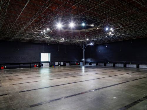 A imagem mostra um amplo pavilhão interno vazio, com piso de concreto liso e marcações escuras retangulares no chão. As paredes laterais são altas e escuras, e no fundo há uma grande porta dupla de vidro, parcialmente iluminada pela luz externa. O teto é sustentado por uma estrutura metálica aparente, composta por treliças e vigas, com luminárias suspensas e refletores ligados, concentrados em um círculo central. Na parte inferior das paredes há várias portas de acesso, caixas de energia e coletores de lixo
