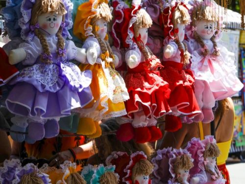 Barraca de feira com várias bonecas de pano coloridas penduradas. Elas têm vestidos volumosos em cores como roxo, amarelo, vermelho, rosa e verde, chapéus combinando e cabelos de lã loira com tranças. O toldo listrado azul e branco cobre a cena.