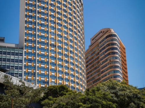 Prédios altos em destaque contra o céu azul. À esquerda, edifício curvo com janelas azuis e detalhes amarelos; à direita, outro prédio laranja com linhas horizontais. Na frente, árvores verdes e barracas de feira com toldos listrados azul e branco. Pessoas circulam.