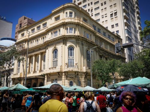 Prédio histórico de cor clara com janelas altas e detalhes arquitetônicos clássicos ocupa o centro da imagem. À frente, feira de rua com barracas de toldos verdes e brancos listrados, rodeada por muitas pessoas caminhando sob o sol.