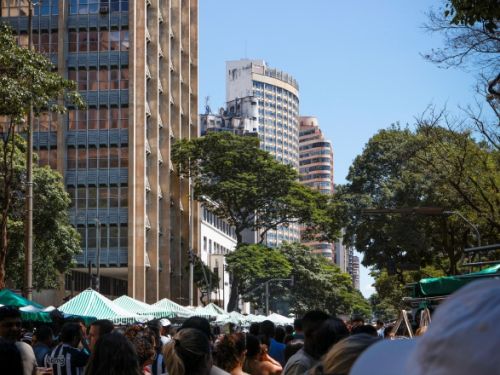 Rua movimentada com muitas pessoas caminhando entre barracas de feira com toldos verdes e brancos listrados. Ao fundo, prédios altos cercados por árvores. O céu está limpo e azul, iluminando a cena urbana.