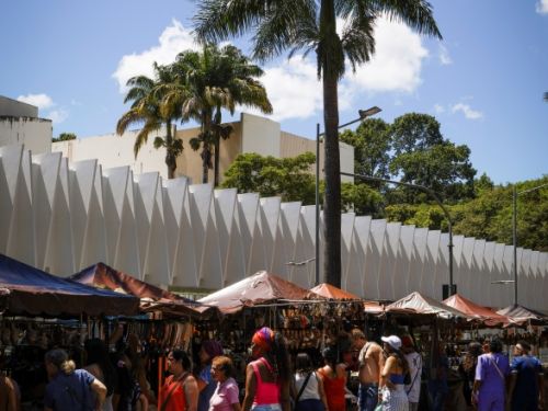 Rua com barracas de feira cobertas por lonas coloridas, onde pessoas observam e compram produtos. Ao fundo, prédio branco de arquitetura moderna com formas triangulares na fachada. Duas palmeiras altas se destacam contra o céu azul com algumas nuvens.
