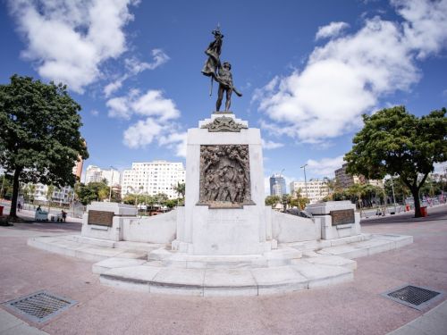 Vista em contra-plongée de um monumento de base de pedra branca e estátuas de bronze. No topo, uma figura masculina segura uma bandeira. O pedestal tem um baixo-relevo detalhado. Ao fundo, árvores, edifícios da cidade e um céu azul com nuvens brancas, fotografado com uma lente grande-angular.