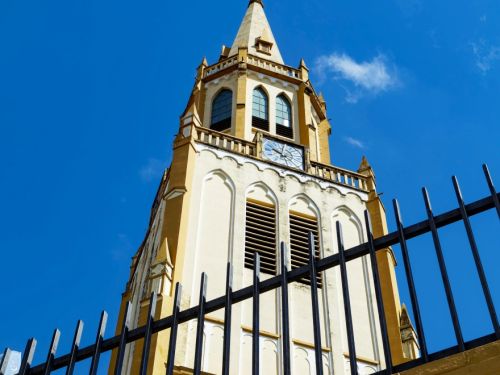 Torre de igreja em tons de amarelo e branco. No topo, há uma cruz branca, janelas ogivais e um relógio. Grades verticais pretas aparecem na parte inferior da imagem. O céu está azul com poucas nuvens.