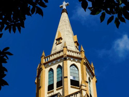 Vista da torre da igreja com cruz no topo, janelas ogivais e relógio visível. A estrutura está emoldurada por galhos escuros na parte superior da imagem, contrastando com o céu azul intenso.