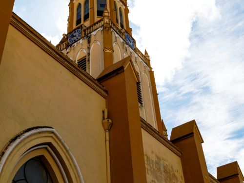 Vista lateral da torre da igreja com céu com nuvens brancas ao fundo. A parede amarelada mostra sinais de desgaste, com janelas góticas e o relógio azul visível no alto da torre.