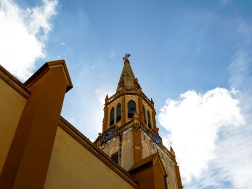 A torre da igreja se projeta sobre a lateral do edifício. O céu azul com nuvens brancas serve de fundo, e vemos o relógio azul nas laterais da torre, que é adornada com janelas ogivais e termina em uma cruz.