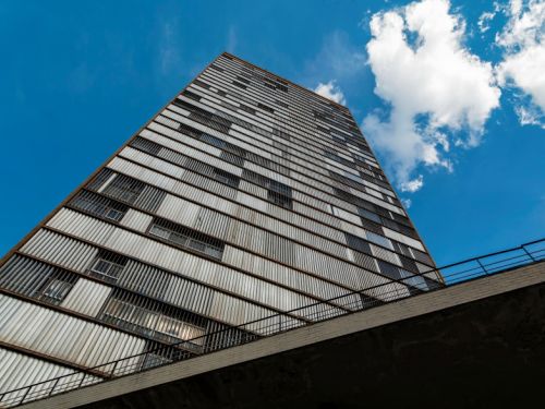 Imagem de baixo para cima de um edifício modernista alto, com fachada marcada por fileiras de janelas e brises verticais e horizontais metálicos, que criam um padrão geométrico repetitivo. A construção ocupa a maior parte da imagem, destacando-se contra o céu azul com algumas nuvens brancas.