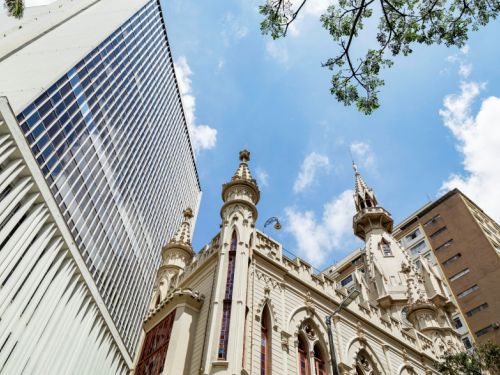 Edifício histórico em estilo gótico, com janelas altas em arco e torres pontiagudas ornamentadas, visto de baixo. Ao fundo, aparecem prédios modernos e o céu azul com nuvens. Ramos de árvore entram pela parte superior da imagem.