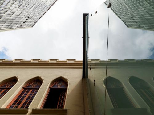 Fachada de prédio histórico com janelas altas em arco e molduras detalhadas. Uma delas está aberta. Ao lado, parede de vidro reflete o edifício. Acima, aparecem arranha-céus modernos e parte do céu nublado.