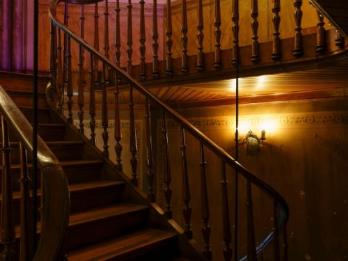 Escada de madeira escura em formato curvo, com corrimão e balaústres torneados. A iluminação amarelada de uma arandela na parede cria um clima aconchegante. No andar superior, paredes claras recebem luz suave em tom lilás, destacando os detalhes clássicos do espaço.