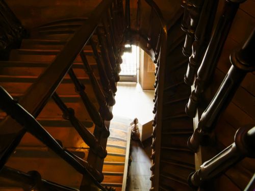 Vista de cima de uma escada de madeira em caracol, com corrimãos grossos e balaústres torneados. A luz natural entra por uma janela ao fundo, iluminando o piso inferior. O contraste entre sombras e reflexos dá profundidade ao espaço, revelando a curvatura elegante da estrutura.