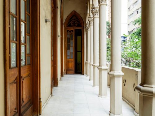 Corredor com piso de mármore claro, ladeado por portas de madeira envidraçadas à esquerda e colunas altas à direita. Os topos das colunas têm detalhes decorativos em relevo, com formas arredondadas e esculpidas. O teto é branco com molduras discretas, e ao fundo há uma porta em arco ogival. Ao lado, vê-se parte de um prédio e árvores.