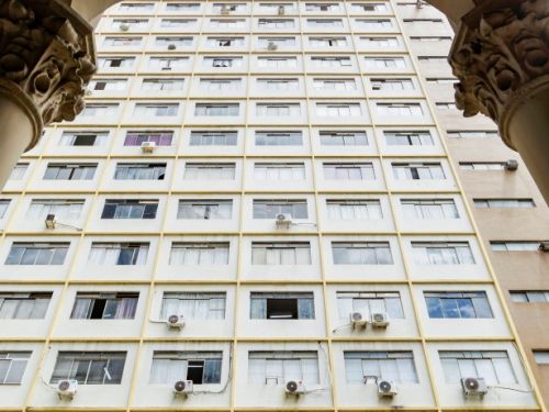 A imagem mostra a vista de um prédio alto e reto, com muitas janelas enfileiradas, algumas abertas e outras com aparelhos de ar-condicionado. Em primeiro plano, aparecem colunas e arcos ornamentados do edifício histórico, criando contraste entre a arquitetura antiga e a moderna.