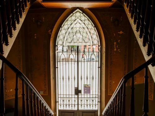Escada de madeira se divide em dois lances curvos que levam a uma porta de ferro alta, em arco ogival, com vitral decorado na parte superior. A luz externa entra pelo vidro, iluminando suavemente as paredes internas de tom terroso e detalhes ornamentais.