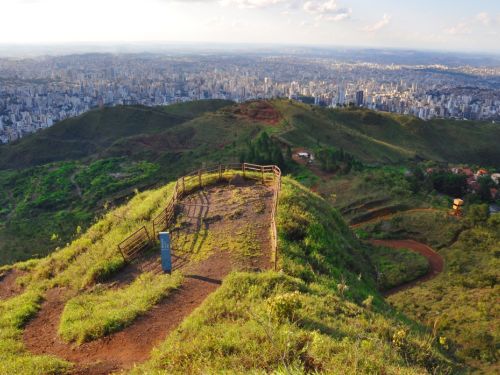 Vista panorâmica da cidade de Belo Horizonte, tirada do topo de um morro, no Parque Serra do Curral. Em primeiro plano, há um mirante circular com uma cerca de madeira e um caminho de terra que leva até ele. O morro é coberto por grama verde. Ao fundo, a vasta paisagem da cidade se estende até o horizonte. O céu está azul com nuvens brancas e esparsas.