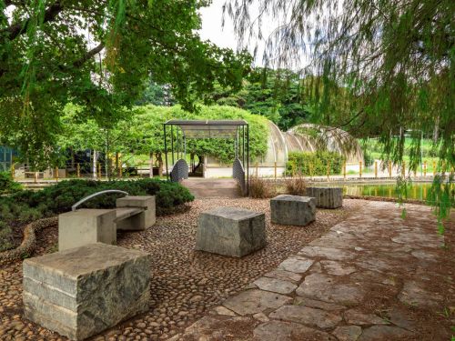 Área de descanso com bancos de concreto e pedras irregulares no chão. Há um pequeno lago ao fundo, com uma ponte metálica conectando ambos os lados. A vegetação ao redor é densa, com árvores altas e plantas rasteiras. O céu está parcialmente nublado.