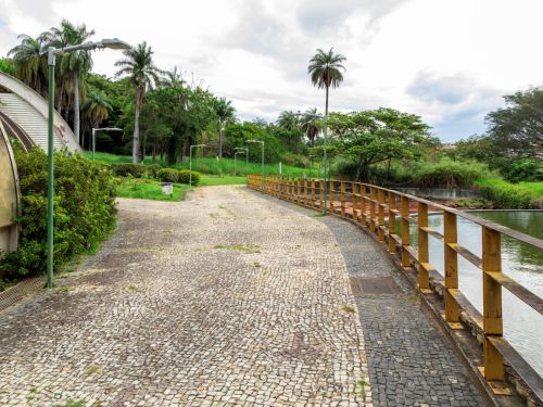 aminho pavimentado com pedras irregulares, acompanhado de uma cerca de madeira baixa e postes de luz ao longo do percurso. A vegetação ao redor é densa, com árvores e arbustos, criando um ambiente tranquilo e sombreado.