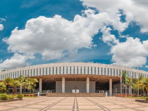 Edifício circular de fachada clara com várias colunas frontais, situado ao fundo de ampla praça com piso geométrico em bege e marrom. Ao lado, palmeiras e árvores. No canto esquerdo aparece um prédio alto marrom, e outros edifícios surgem à direita sob céu azul com nuvens.