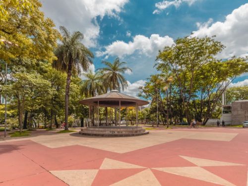 Coreto circular com cobertura metálica no centro de praça arborizada. Ao redor, árvores altas e palmeiras. O piso tem desenhos geométricos em vermelho e bege. Ao fundo, pessoas caminham e bicicletas circulam. O céu está azul com nuvens brancas.