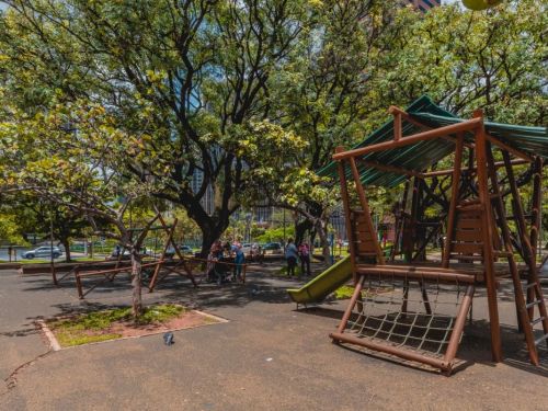 Parque arborizado com brinquedos de madeira. À direita, estrutura com escada, escorregador verde e rede de cordas. Pessoas estão reunidas ao fundo sob as árvores. O chão é pavimentado em asfalto escuro, com canteiros pequenos. Árvores de copa densa ocupam toda a área.