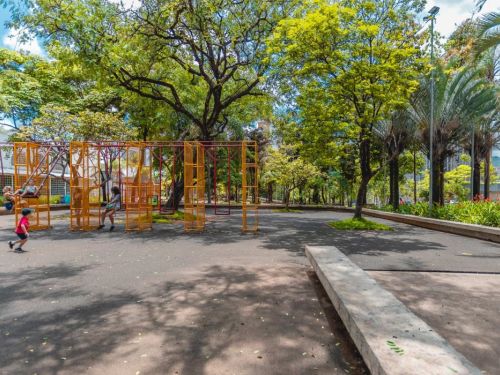 Playground ao ar livre com estruturas metálicas de cor laranja e balanços vermelhos. Crianças brincam no local. Árvores altas com copas verdes fazem sombra sobre o piso escuro. Ao fundo, caminhos pavimentados e mais áreas verdes delimitam o espaço.