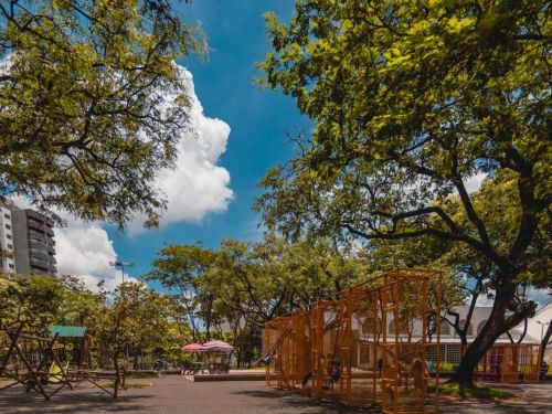 Praça arborizada em dia ensolarado. À direita, brinquedos metálicos laranja com balanços. À esquerda, brinquedos de madeira. Pessoas circulam pela área sombreada por árvores. No centro, quiosque com guarda-sóis coloridos. Ao fundo, edifícios altos são visíveis.