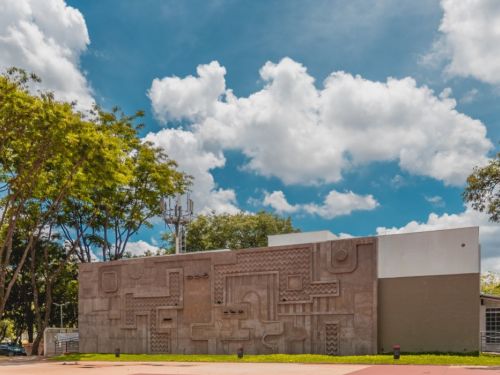 Parede grande de concreto cinza ocupa o centro da imagem, com relevos geométricos de linhas, curvas e padrões em zigue-zague. À esquerda, árvores altas com folhagem verde. O céu está azul com nuvens brancas espalhadas. O piso da praça em primeiro plano é avermelhado.