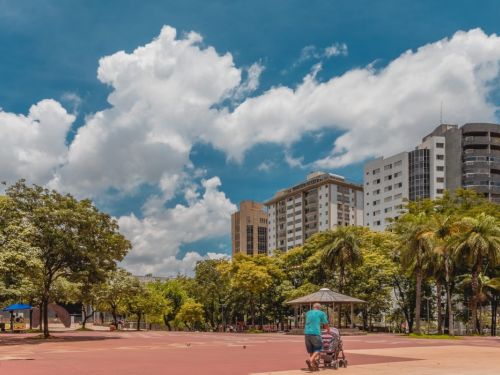 Ampla praça com piso em tons de vermelho e bege, pessoas caminhando e edifícios altos ao fundo. À esquerda, criança de roupa vermelha. No centro, homem empurra carrinho de bebê. Muitas árvores e palmeiras margeiam o espaço, sob céu azul com nuvens brancas volumosas.