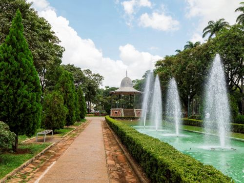 Fonte em lago retangular verde, com jatos de água alinhados simetricamente. Cercada por arbustos baixos, árvores e caminhos de tijolos. Coreto metálico de teto cinza surge ao fundo. Bancos laterais completam o espaço de convivência.